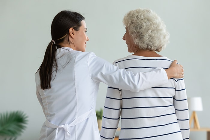 Rear back view of young female doctor physiotherapist caretaker help injured old adult grandma patient assisting holding walking with senior lady at home hospital